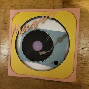 May include: A layered paper art piece featuring a retro turntable design. The artwork has a black vinyl record with a purple center, a light blue turntable, and the word "Music" in pink script. The piece is framed in yellow and pink.