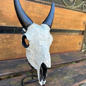 May include: A decorative animal skull sculpture featuring a white mosaic tile surface on the cranium and smooth black horns. The skull is mounted on a circular black metal stand, showcasing a high-contrast design with black accents in the eye sockets.