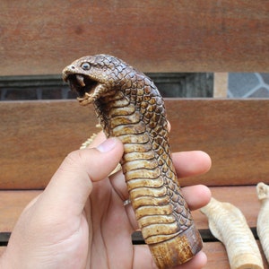 Può includere: Un'intagliatura dettagliata di una testa e di un corpo di cobra, tenuta in mano. Il serpente è marrone e beige con la bocca aperta, che rivela i denti. Il corpo ha un motivo a squame. Ci sono altri tre pezzi intagliati sullo sfondo.