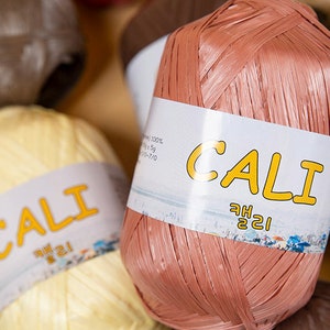 May include: Close-up of a pink ball of yarn with a white label that reads "CALI" in yellow letters. The yarn is made of thin, woven strands and has a slightly textured surface.