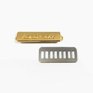 Puede incluir: Dos piezas de ferretería metálicas. Una etiqueta rectangular dorada con la palabra "handmade" en escritura cursiva. Debajo, una pieza rectangular plateada con siete recortes rectangulares. Ambas piezas están sobre un fondo blanco.