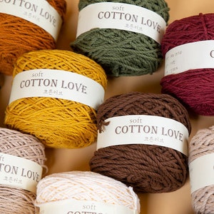 Puede incluir: Una colecci&oacute;n de ovillos de lana en varios colores, incluyendo amarillo mostaza, rojo oscuro y verde oliva. Cada ovillo est&aacute; etiquetado con "soft COTTON LOVE" en ingl&eacute;s y coreano. La lana est&aacute; bien enrollada, lo que sugiere que es adecuada para proyectos de tejido o ganchillo.