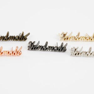 Puede incluir: Una colección de dijes metálicos con la palabra "Handmade" en varios acabados: bronce, oro rosa, negro, dorado y plateado. Los dijes están en una fuente cursiva y tienen pequeños postes para sujetar, adecuados para joyería o manualidades.