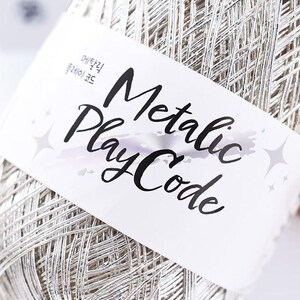May include: A close-up of a spool of silver metallic yarn. The yarn is wrapped around itself, and a white label with the words "Metallic Play Code" in black script is wrapped around the yarn.