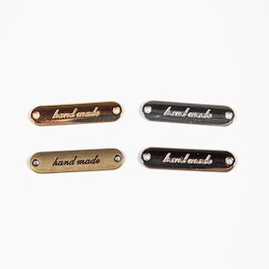 Peut inclure: Quatre étiquettes métalliques avec les mots "hand made" gravés. Deux étiquettes sont dorées et deux sont argentées. Chaque étiquette comporte deux petits trous à chaque extrémité. Les étiquettes sont de forme ovale et sur fond blanc.
