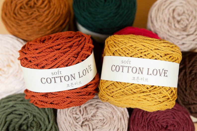 Puede incluir: Primer plano de varios ovillos de hilo de algod&oacute;n suave en varios colores, incluyendo &oacute;xido, amarillo mostaza y verde. Cada ovillo tiene una etiqueta blanca con el texto "soft COTTON LOVE" y caracteres coreanos. Ideal para tejer y hacer ganchillo.