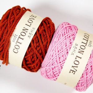 Puede incluir: Dos ovillos de hilo de algod&oacute;n suave en tonos &oacute;xido y rosa. Cada ovillo tiene una etiqueta que dice "COTTON LOVE" en ingl&eacute;s y coreano. El ovillo &oacute;xido est&aacute; etiquetado "36 합" y el rosa "18 합".