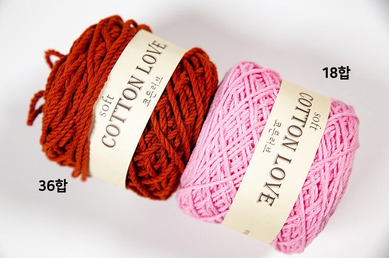 Puede incluir: Dos ovillos de hilo de algod&oacute;n suave en tonos &oacute;xido y rosa. Cada ovillo tiene una etiqueta que dice "COTTON LOVE" en ingl&eacute;s y coreano. El ovillo &oacute;xido est&aacute; etiquetado "36 합" y el rosa "18 합".