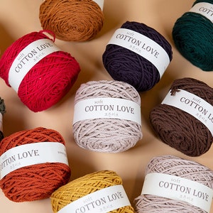 Puede incluir: Una colecci&oacute;n de ovillos de lana coloridos en varios tonos, incluyendo rojo, verde, marr&oacute;n y amarillo. Cada ovillo est&aacute; etiquetado con "Soft Cotton Love" en texto blanco. Los ovillos est&aacute;n dispuestos sobre un fondo neutro.