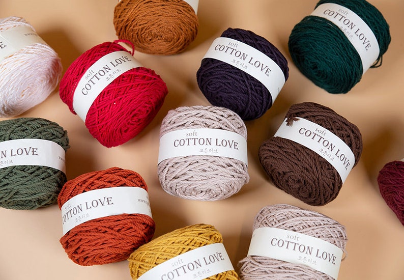 Puede incluir: Una colecci&oacute;n de ovillos de lana coloridos en varios tonos, incluyendo rojo, verde, marr&oacute;n y amarillo. Cada ovillo est&aacute; etiquetado con "Soft Cotton Love" en texto blanco. Los ovillos est&aacute;n dispuestos sobre un fondo neutro.