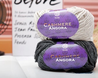 May include: Two skeins of yarn stacked vertically, one light beige and one dark charcoal gray. Each skein features a purple paper label with the text YEONIL and CASHMERE ANGORA printed in gold lettering, along with a circular logo at the top.