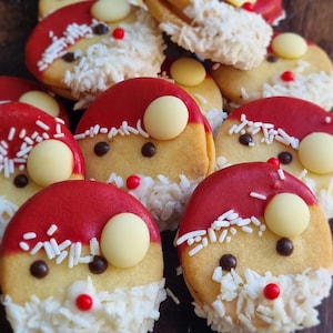 Op de afbeelding: Ronde koekjes versierd als gezichten van de Kerstman. Elk koekje heeft een rode glazuurmuts, een witte chocoladeknop, witte hagelslag, twee donkere chocoladeogen, een rode snoepneus en een baard gemaakt van geraspte kokosvlokken.
