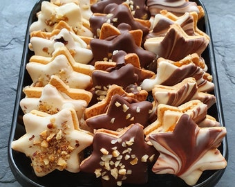 May include: A black tray filled with star-shaped biscuits. Some are dipped in white chocolate and topped with nuts, others coated in dark chocolate or swirled with chocolate. The biscuits are arranged in neat rows, a festive treat.