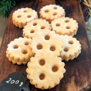 Könnte beinhalten: Nahaufnahme eines Stapels hausgemachter Kekse in hellbrauner Farbe mit einer Bestäubung von Puderzucker. Die Kekse haben ein dekoratives Design mit vier Löchern in jedem Keks. Die Kekse sind auf einer Holzoberfläche angeordnet.