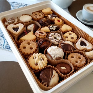Può includere: Una scatola bianca piena di una varietà di biscotti, tra cui biscotti al cioccolato, alla vaniglia e biscotti ripieni di frutta. I biscotti sono disposti in un motivo decorativo e sono pronti per essere gustati.