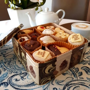 Op de afbeelding: Een bruine en gouden blikken doos gevuld met verschillende koekjes. De koekjes zijn in een rasterpatroon gerangschikt en bevatten chocolade-, vanille- en fruitsmaken. De doos heeft een deksel met een cupcake-ontwerp en de tekst "Lebe den Moment" aan de voorkant.