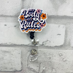 May include: A white badge reel with a black retractable cord. The badge holder features a colorful graphic with the text "My Body My Rules" in a retro font with flowers.