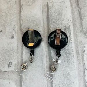 Peut inclure: Deux porte-badges r&eacute;tractables noirs avec des clips argent&eacute;s et des sangles en plastique transparent. Les porte-badges sont sur un fond de briques blanches.