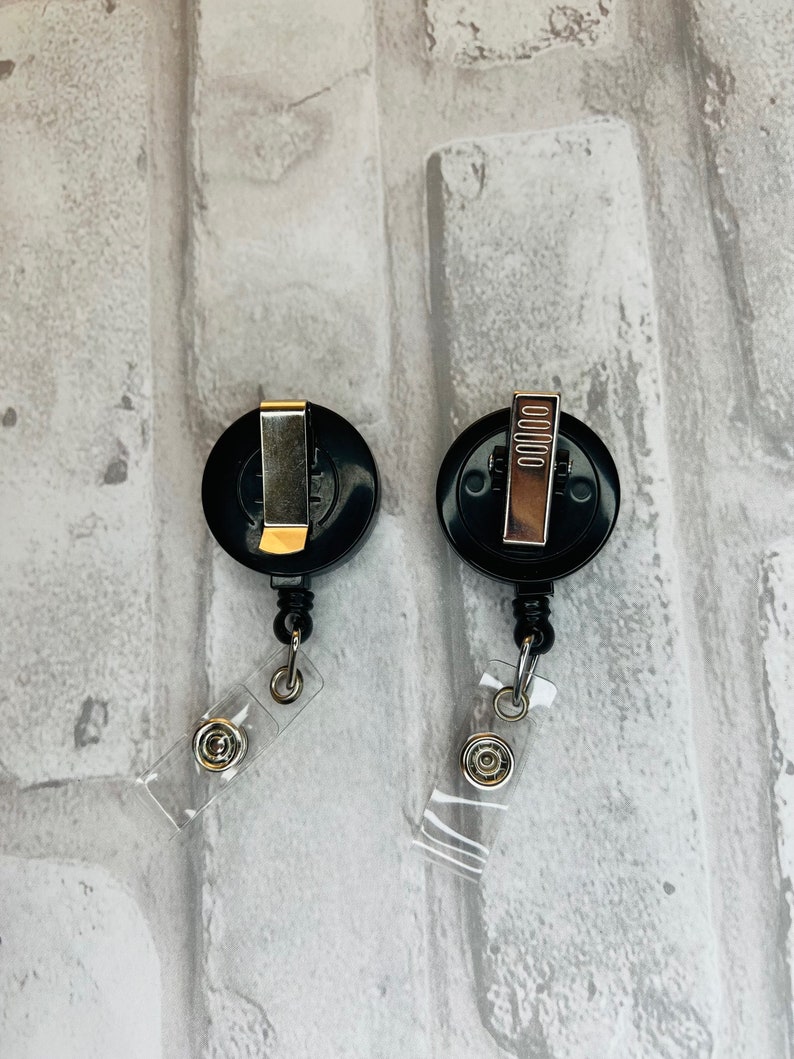 Peut inclure: Deux porte-badges r&eacute;tractables noirs avec des clips argent&eacute;s et des sangles en plastique transparent. Les porte-badges sont sur un fond de briques blanches.
