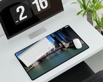 May include: A rectangular mousepad featuring a cityscape with a river and tall buildings. A white keyboard and mouse are on the mousepad. A computer monitor displays the time as 2:16 PM. The setup is on a white desk.