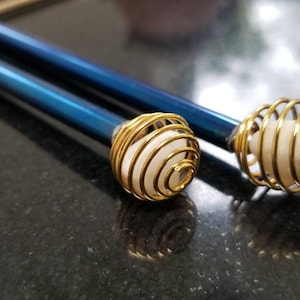 Puede incluir: Dos varillas metálicas azules con extremos de jaula de alambre dorado que contienen esferas blancas. Las esferas están encerradas en las jaulas de alambre.