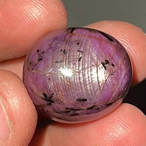 Può includere: Una grande pietra preziosa ovale di colore viola con inclusioni nere. La pietra preziosa è liscia e lucidata.