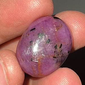May include: A smooth, oval-shaped, purple gemstone with black inclusions. The gemstone is polished and has a glossy finish.