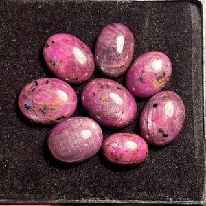 Può includere: Nove pietre preziose rosa di forma ovale con inclusioni nere. Le pietre preziose sono disposte a forma di fiore su una superficie di velluto nero.