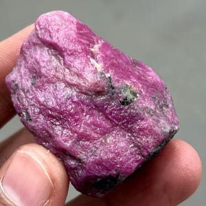 Könnte beinhalten: Ein roher, ungeschliffener Rubin-Edelstein. Der Rubin hat eine tiefe, lebendige rosa-rote Farbe mit einer strukturierten Oberfläche. Kleine schwarze Einschlüsse sind sichtbar. Der Edelstein wird in einer Hand gehalten.