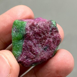 Könnte beinhalten: Ein roher Rubin-Edelstein mit tiefroter und grüner Farbe. Der Rubin wird in einer Hand gehalten und zeigt seine natürliche, unpolierte Textur. Der Stein hat eine raue, unregelmäßige Form, die seine natürliche Schönheit hervorhebt.