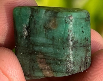 May include: A dark green emerald gemstone with black inclusions. The emerald is cut into a rectangular shape and has a smooth, polished surface.
