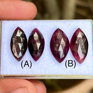 May include: Four marquise-cut gemstones in a clear plastic case. The gems are a deep red-purple colour with faceted surfaces. The gems are labelled (A) and (B). The gems are held in a person's hand.