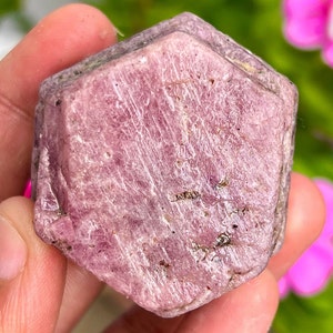 Puede incluir: Un gran cristal de rubí rosa facetado con forma hexagonal. El cristal tiene una textura rugosa y natural y es translúcido.