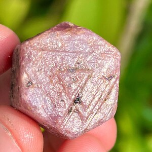 May include: A pink ruby crystal with a hexagonal shape and a shiny, sparkly surface.