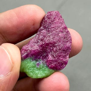 May include: A rough-cut gemstone with a vibrant color contrast. The upper portion displays a deep, rich magenta hue, while the lower section is a bright, verdant green. The stone's surface has a textured, natural appearance.