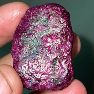 Könnte beinhalten: Ein roher, rosafarbener Rubin mit silbernen und grünen Einschlüssen. Der Edelstein hat eine unregelmäßige Form und eine glänzende, polierte Oberfläche.