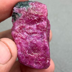 Può includere: Una gemma di rubino grezza e non tagliata. Il rubino è di un colore rosso-rosato intenso e vibrante, con una superficie leggermente strutturata. Una piccola area di verde scuro e nero è visibile nella parte superiore della pietra. La gemma è tenuta in una mano.