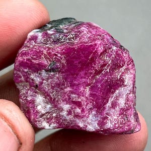 May include: A large, rough, pink ruby crystal with white and black inclusions. The crystal has a natural, unpolished finish.