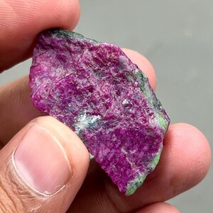 Puede incluir: Una gema de rubí en bruto, de color magenta intenso, con toques de verde y gris. El mineral en bruto presenta una superficie texturizada, mostrando variaciones naturales de color y forma. El rubí se sostiene en una mano.