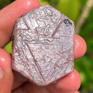 May include: A large, hexagonal, pink ruby crystal with a silvery sheen. The crystal has a rough, natural surface and is held in a hand.