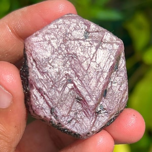 May include: A large, faceted, pink ruby crystal with a metallic sheen. The crystal is shaped like a hexagon and has a rough, natural texture.