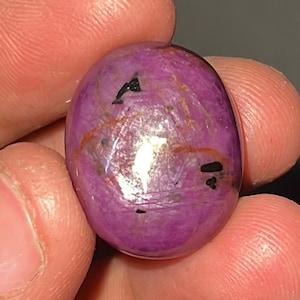 Può includere: Una gemma lucida di forma ovale con una profonda tonalità magenta. La pietra presenta inclusioni scure e sottili venature arancioni sulla sua superficie. La gemma è tenuta tra le dita, mostrando la sua qualità liscia e riflettente.