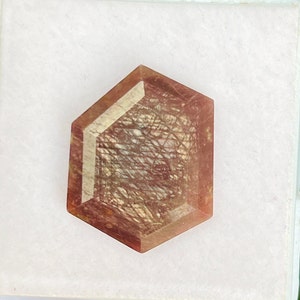 Peut inclure: Une pierre précieuse hexagonale, brun rougeâtre, avec des inclusions internes créant un motif en forme de toile. La pierre facettée est posée sur un fond blanc. La couleur et la clarté de la gemme sont mises en valeur.
