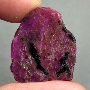 Könnte beinhalten: Ein roher Rubin-Edelstein in tiefem Magenta, mit dunklen schwarzen Einschlüssen. Die Oberfläche hat eine leicht unebene Textur mit Bereichen hellerer und dunklerer Färbung. Der Edelstein wird zwischen Fingern gehalten.