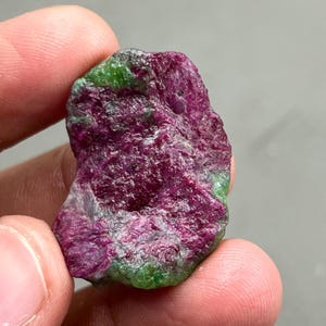 May include: A raw ruby gemstone with a deep magenta hue, accented by patches of vibrant green. The rough, natural texture of the stone is visible, showcasing its unpolished state. The ruby is held between fingers, highlighting its size and shape.
