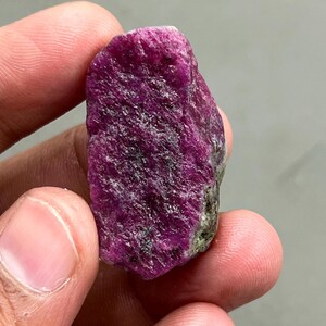 May include: A raw ruby gemstone, displaying a deep magenta colour with visible crystalline structures. The uncut mineral has a textured surface and natural imperfections, highlighting its unrefined state. The ruby is held in a hand, emphasising its size and form.