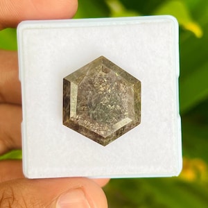 May include: A hexagonal, faceted gemstone with a dark brown and gray color. The gemstone is set in a white, square, velvet-lined box.