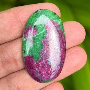 Könnte beinhalten: Ovaler, polierter Edelstein mit lebendigen Farben. Der Stein zeigt eine Mischung aus tiefem Magenta und leuchtend grünen Flecken. Der Edelstein wird in einer Hand gehalten, was seine natürliche Schönheit und einzigartige Farbkombination hervorhebt.