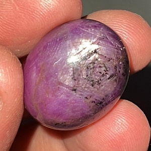Può includere: Una pietra preziosa viola con una forma ovale liscia e un interno scuro e nuvoloso. La pietra ha un'alta lucidatura ed è probabilmente un rubino.