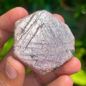 Può includere: Un grande cristallo di rubino rosa grezzo con forma esagonale e lucentezza argentata. Il cristallo ha un aspetto naturale e non levigato.